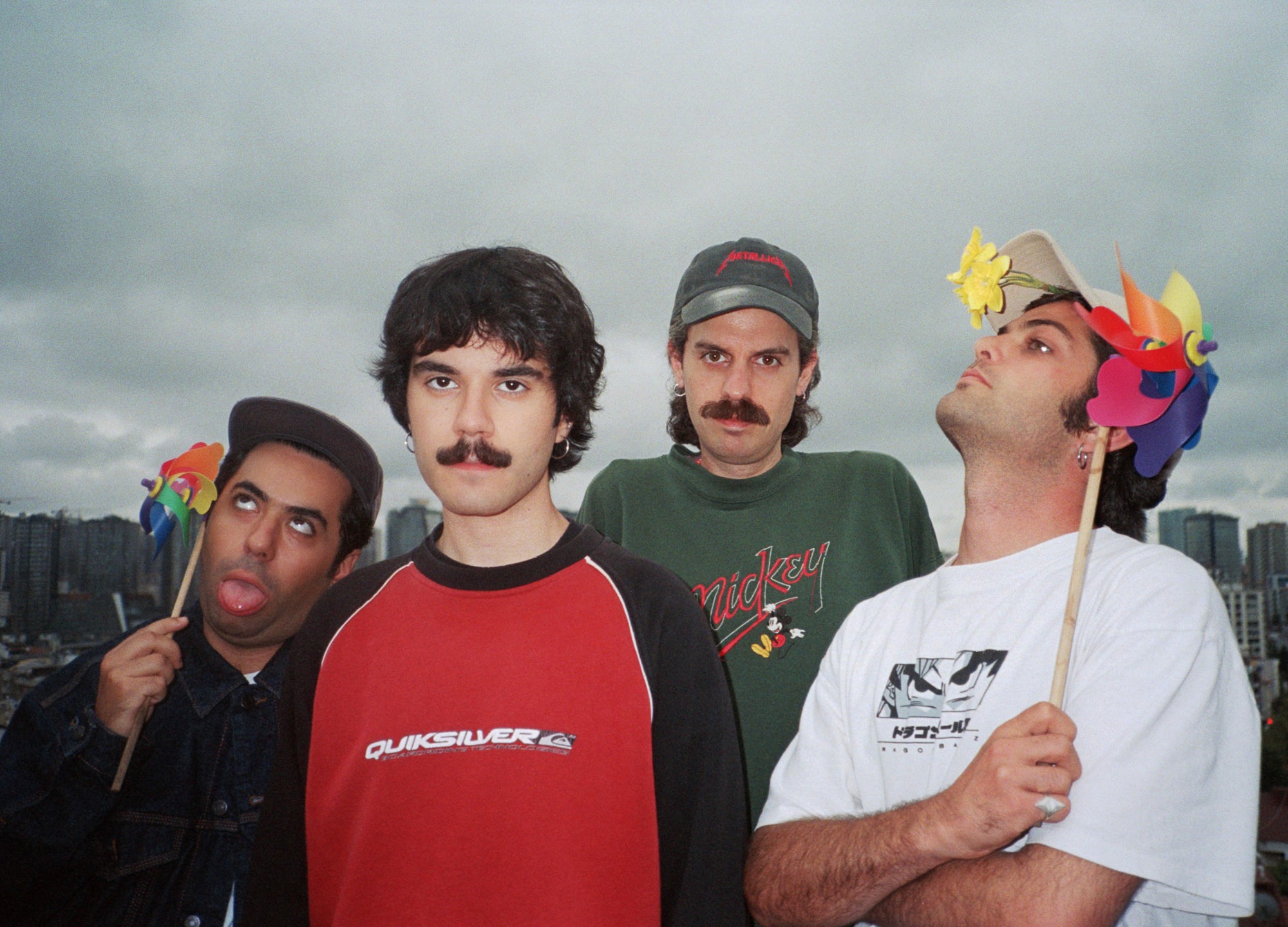 A group of four individuals stands on a rooftop against a cloudy cityscape, each holding colorful pinwheels, with three of the individuals looking directly at the camera and one gazing upward.