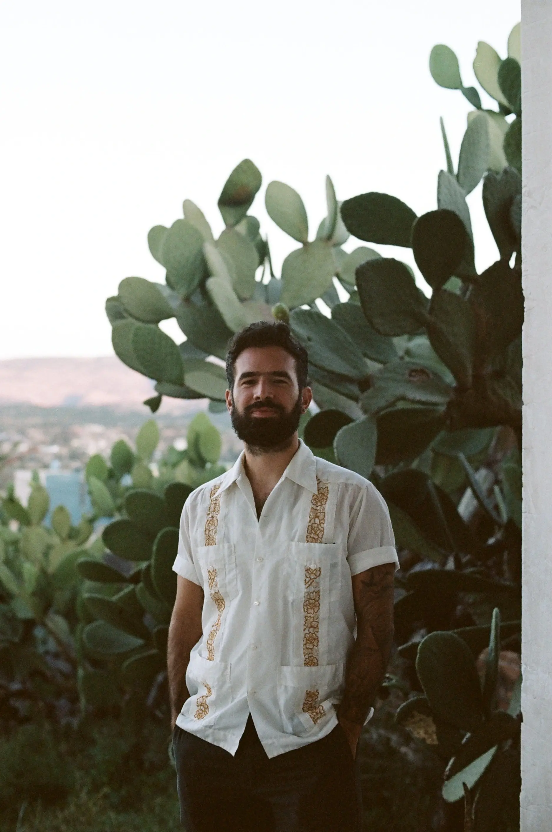 Mario, a bearded individual stands in front of large green cacti, wearing a white shirt with decorative patterns, against a distant backdrop of rolling hills under a clear sky.