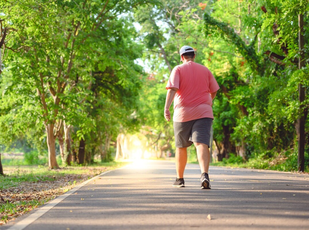 plus-sized man sweating as he walks along a park trail to reach his goal of how many miles per day to lose weight