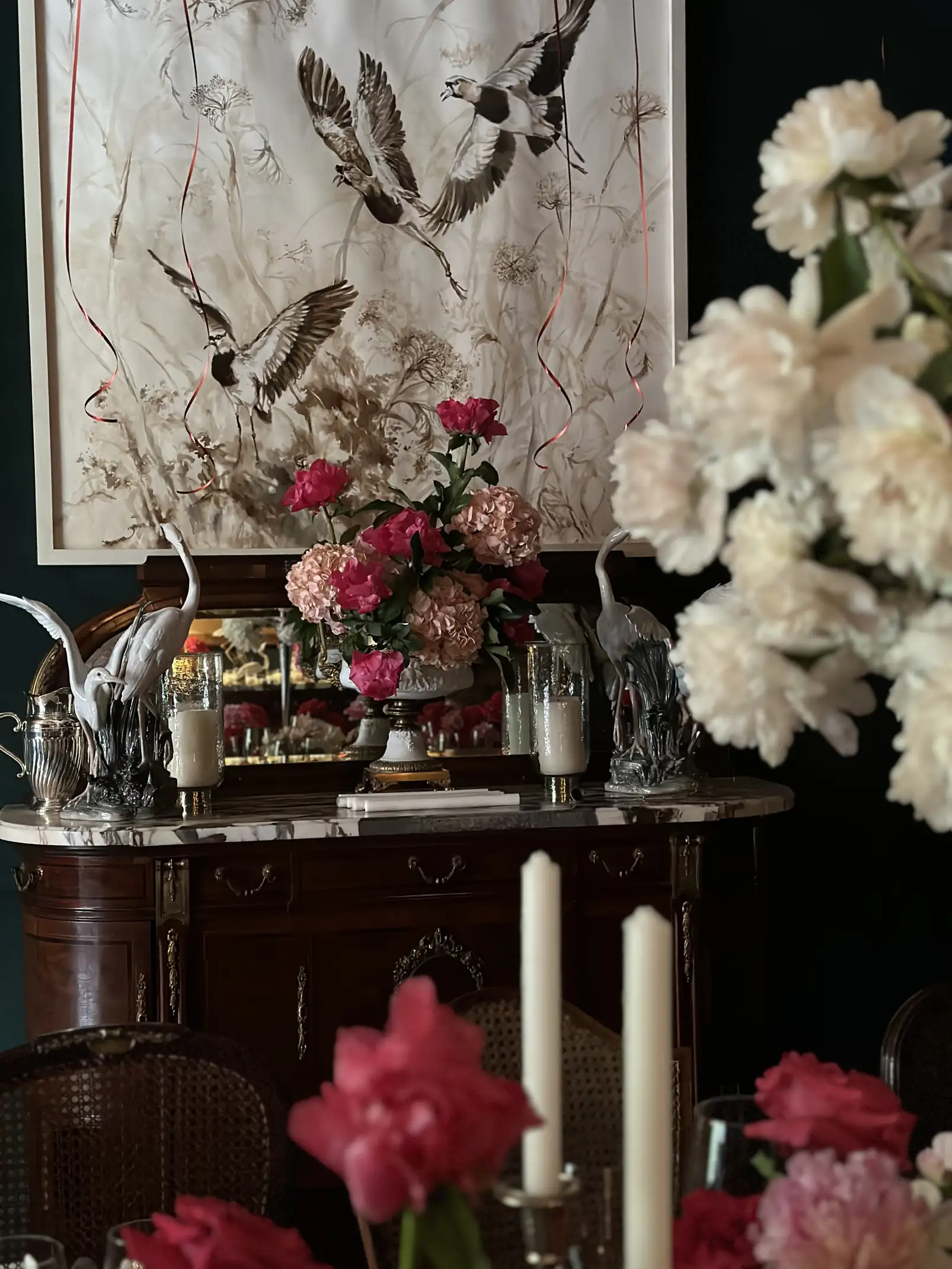 Composición floral con hortensias y rosas en tonos rosados sobre una cómoda de madera con superficie de mármol, acompañada por candelabros y esculturas de aves, enmarcada por una pintura de pájaros en vuelo que aporta un aire clásico y poético al espacio.