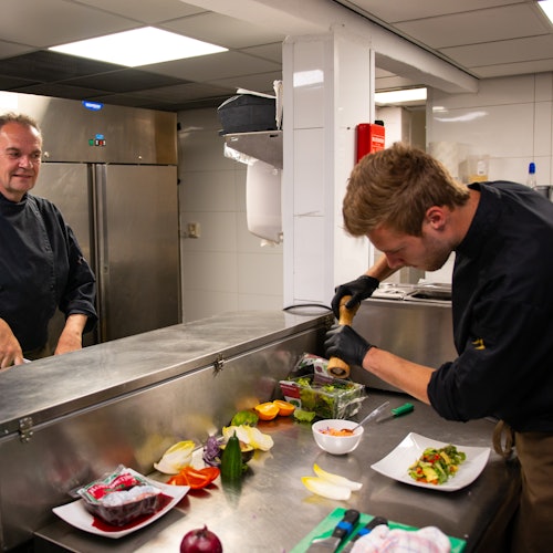 Two chefs cooking in a kitchen with utensils, fresh vegetables, and dishes on the stainless steel counter.