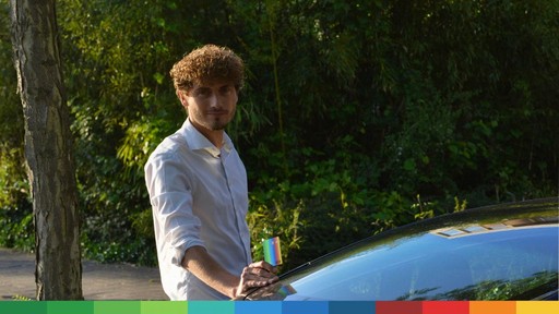 A young man in a light shirt stands near a car, surrounded by greenery in a sunny outdoor setting.