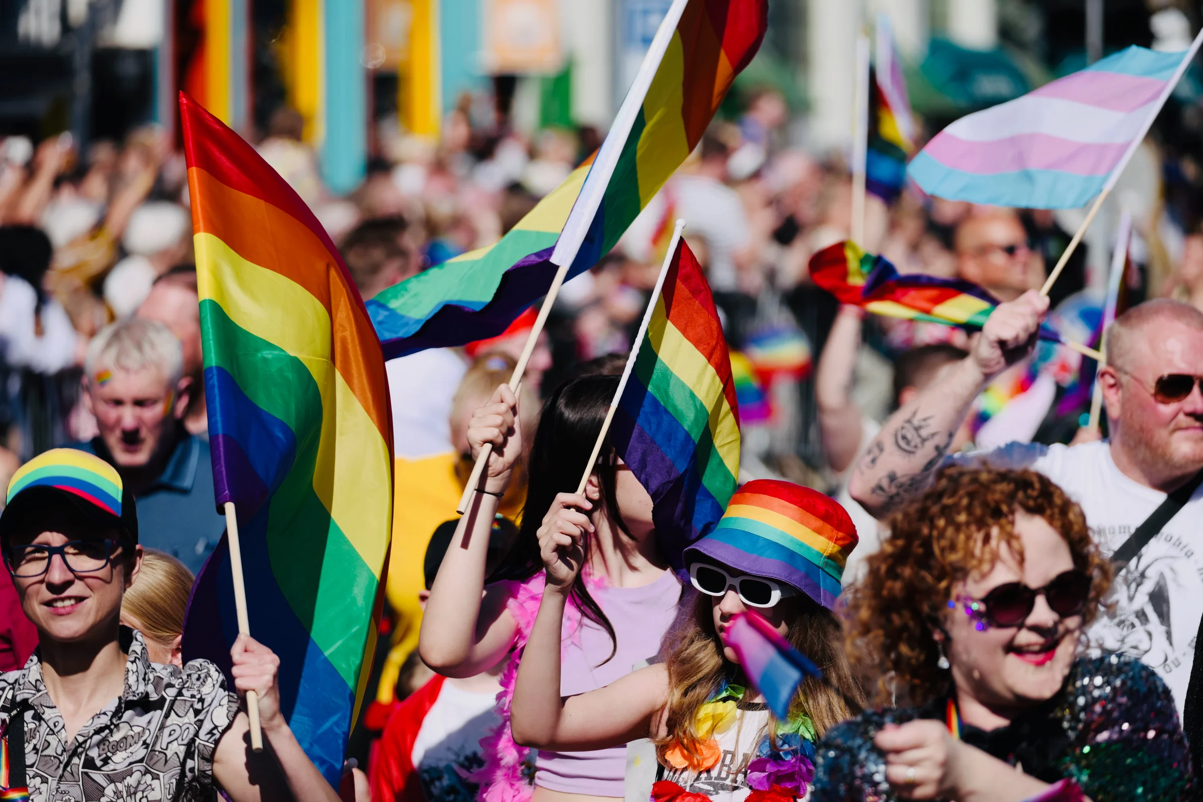 A vibrant crowd of participants waving rainbow flags and a transgender pride flag during a sunny outdoor pride parade.