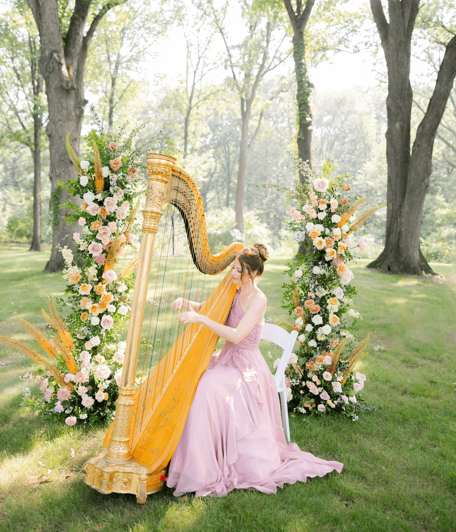 Dawn Bishop playing her harp in a beautiful outdoor setting amungst flowers while wearing a pink gown.