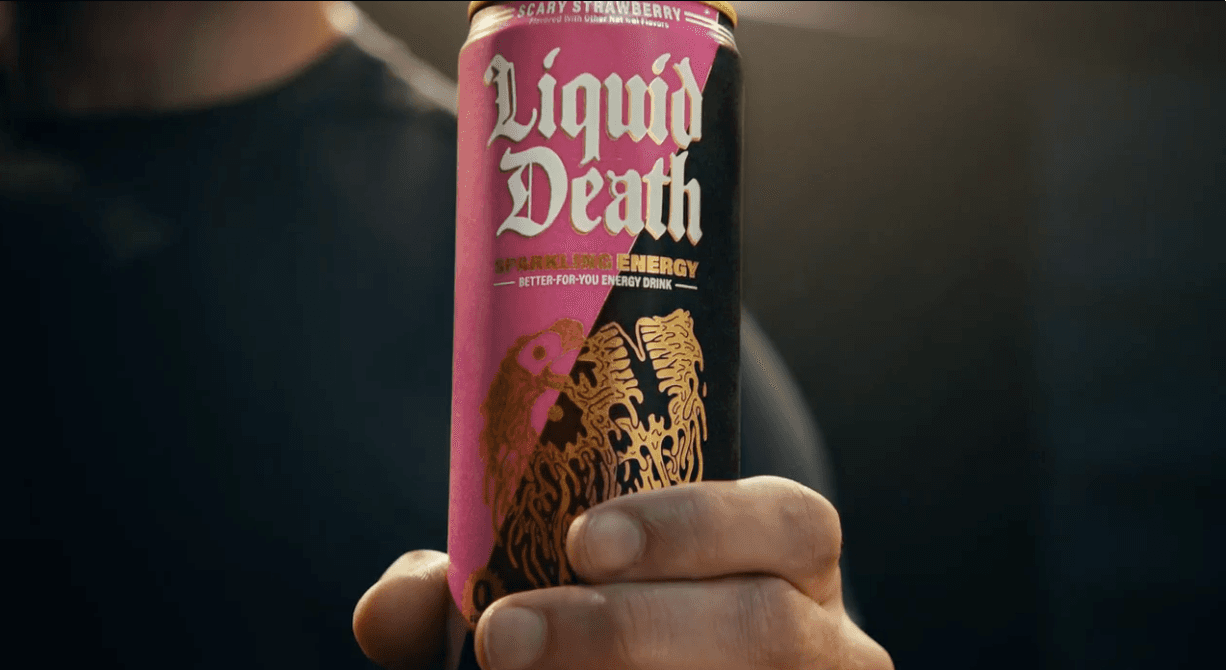 A person's hand holds up a tall aluminum can of Liquid Death Sparkling Energy drink. The can features a split pink and black background with the brand name "Liquid Death" in large, white, gothic font. Below the name, text reads "SPARKLING ENERGY" and "BETTER-FOR-YOU ENERGY DRINK". The top rim of the can indicates the flavor is "SCARY STRAWBERRY". The bottom half of the can displays a stylized, melting golden graphic.