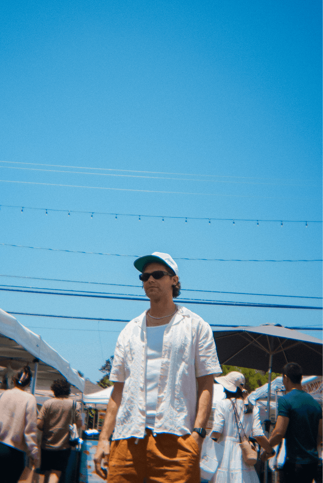 a young man in a white shirt , a baseball hat, and terracotta shorts standing in the middle of a farmers market on a clear bright day