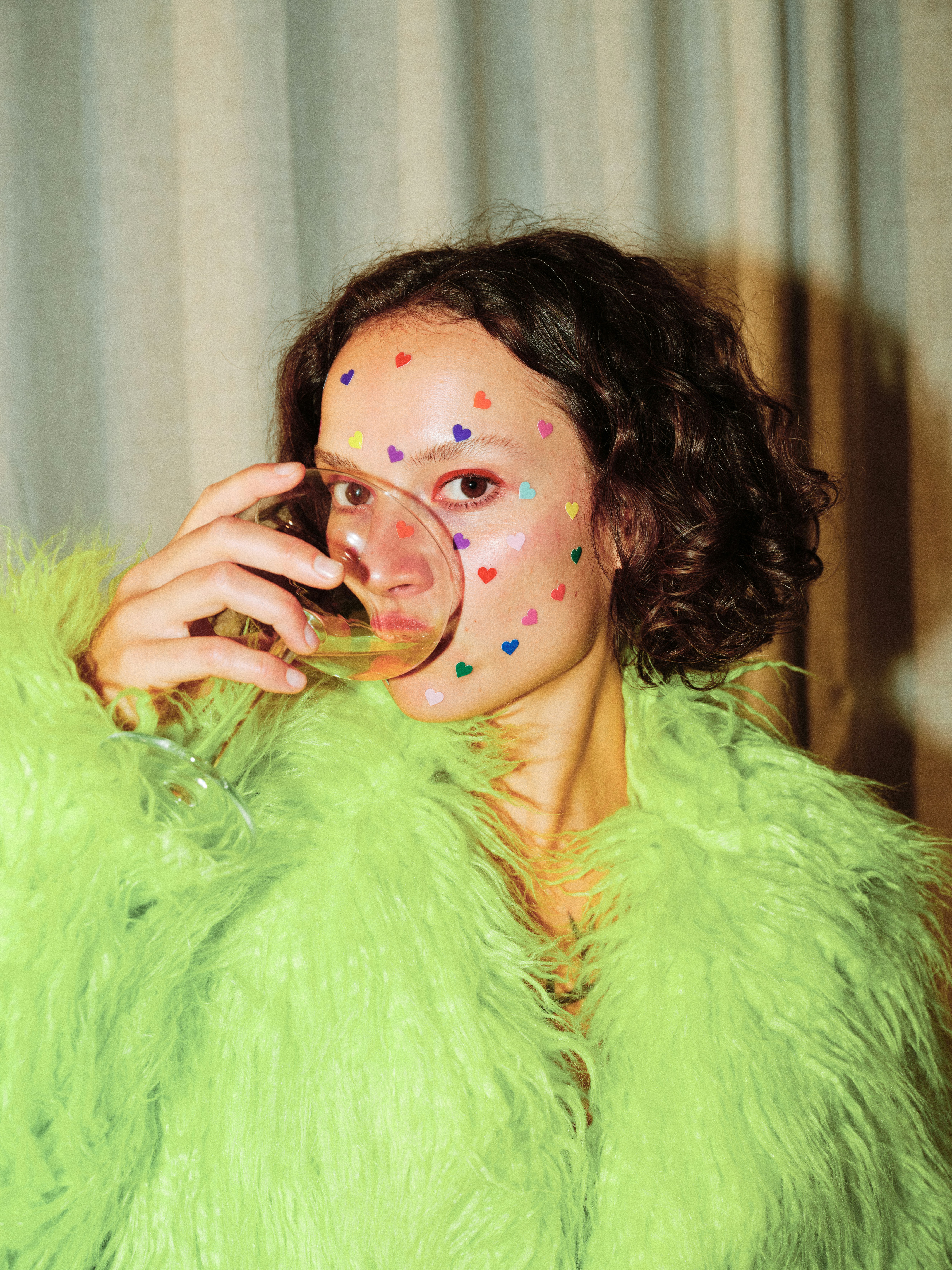 Editorial portrait of a curly-haired model wearing a lime green faux fur coat with multicoloured heart stickers on her face, holding a wine glass