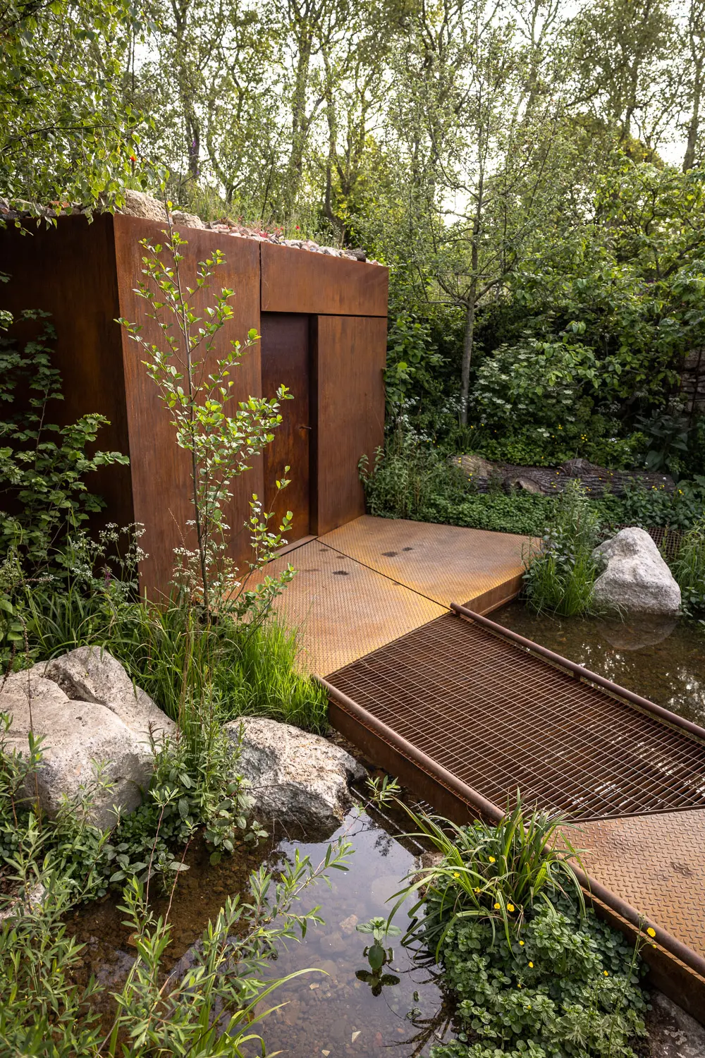 A wooden cabin beside a serene path leads through lush greenery and rocks, with a gentle stream nearby.