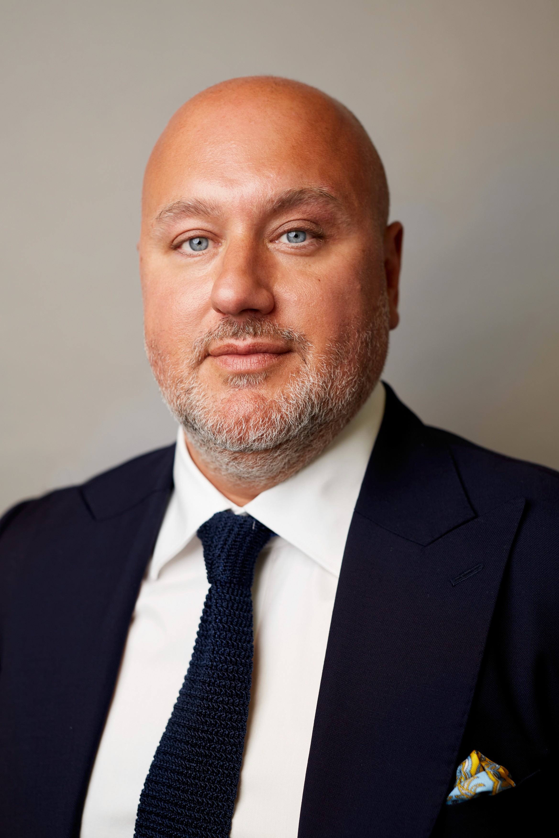 Bald man in a dark suit and blue tie with blue eyes.