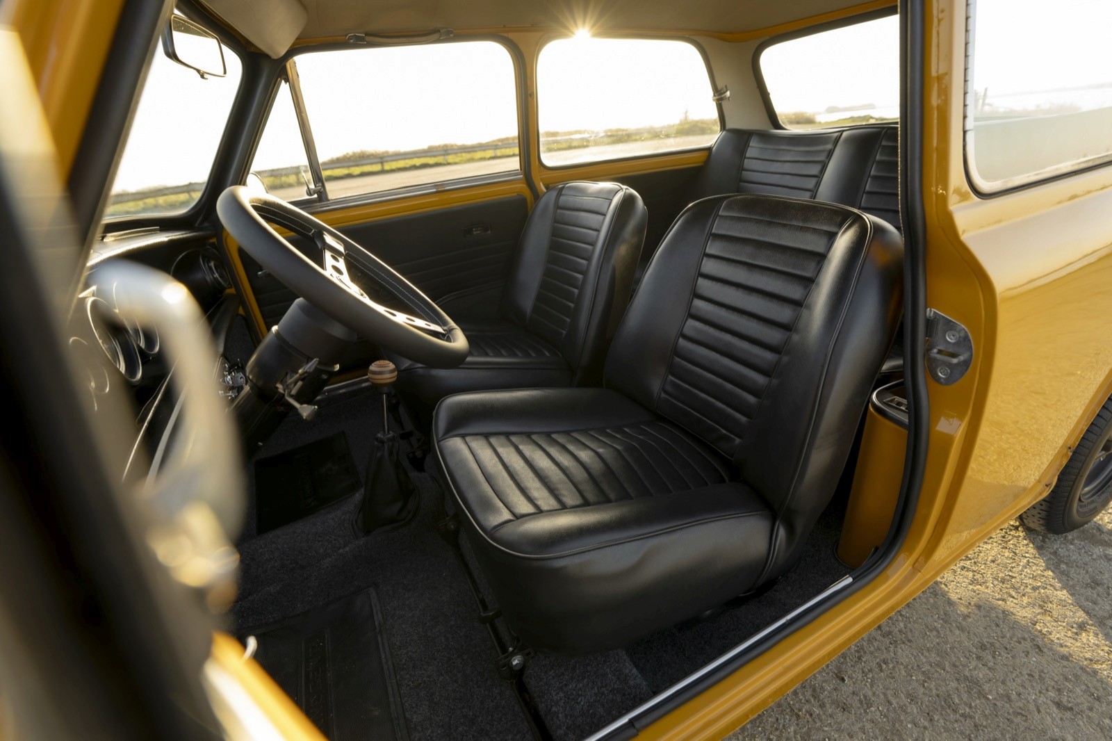 Interior view of a classic car showing black leather seats and steering wheel, with sunlight filtering through windows.