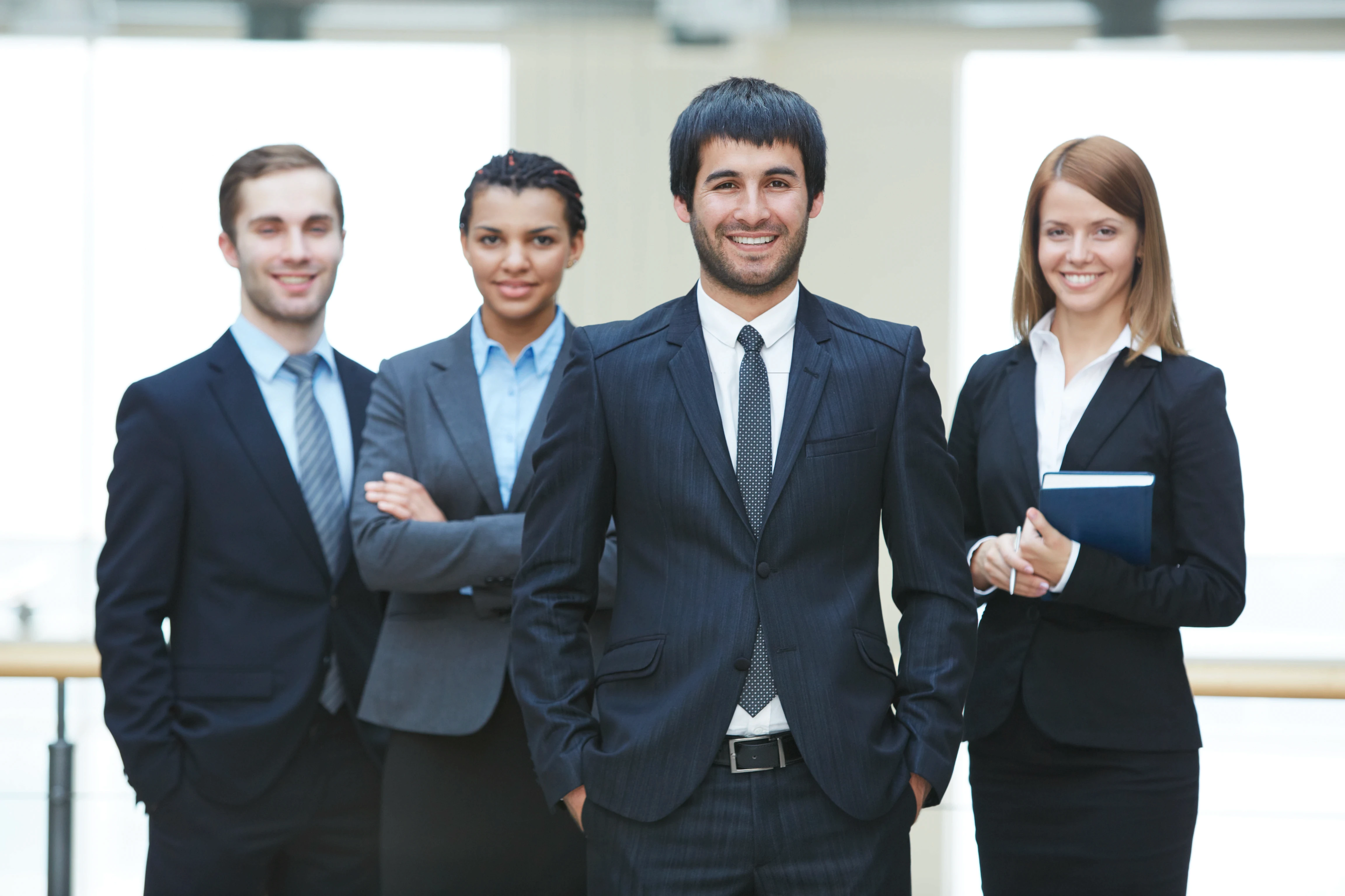 A confident group of four diverse professionals in suits stand smiling in a bright office setting, conveying teamwork and professionalism.