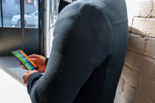 A person in a dark jacket holds a smartphone, looking at it, with a window view in the background.