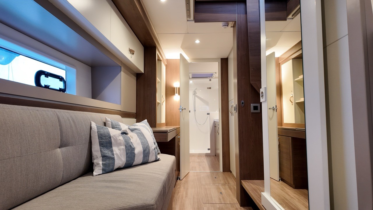 Bali 4.4 sailing catamaran aft hull cabin interior showing a grey upholstered sofa berth with striped cushion, warm oak veneer cabinetry, light wood flooring, hull portlight window, and open en-suite shower compartment with chrome fittings and mirrored bathroom beyond