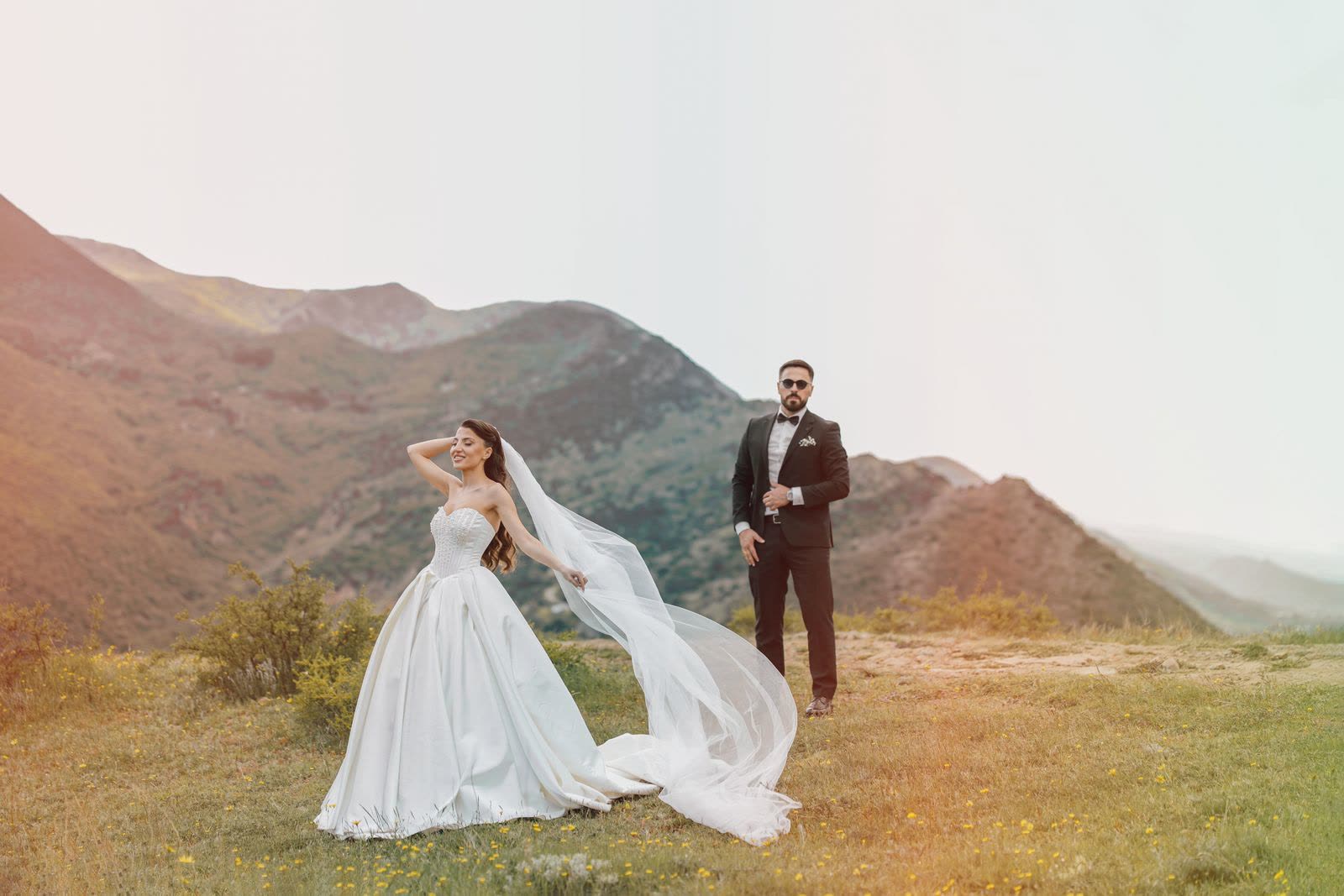 Bride with a long veil beside the groom on a hill with mountains behind them