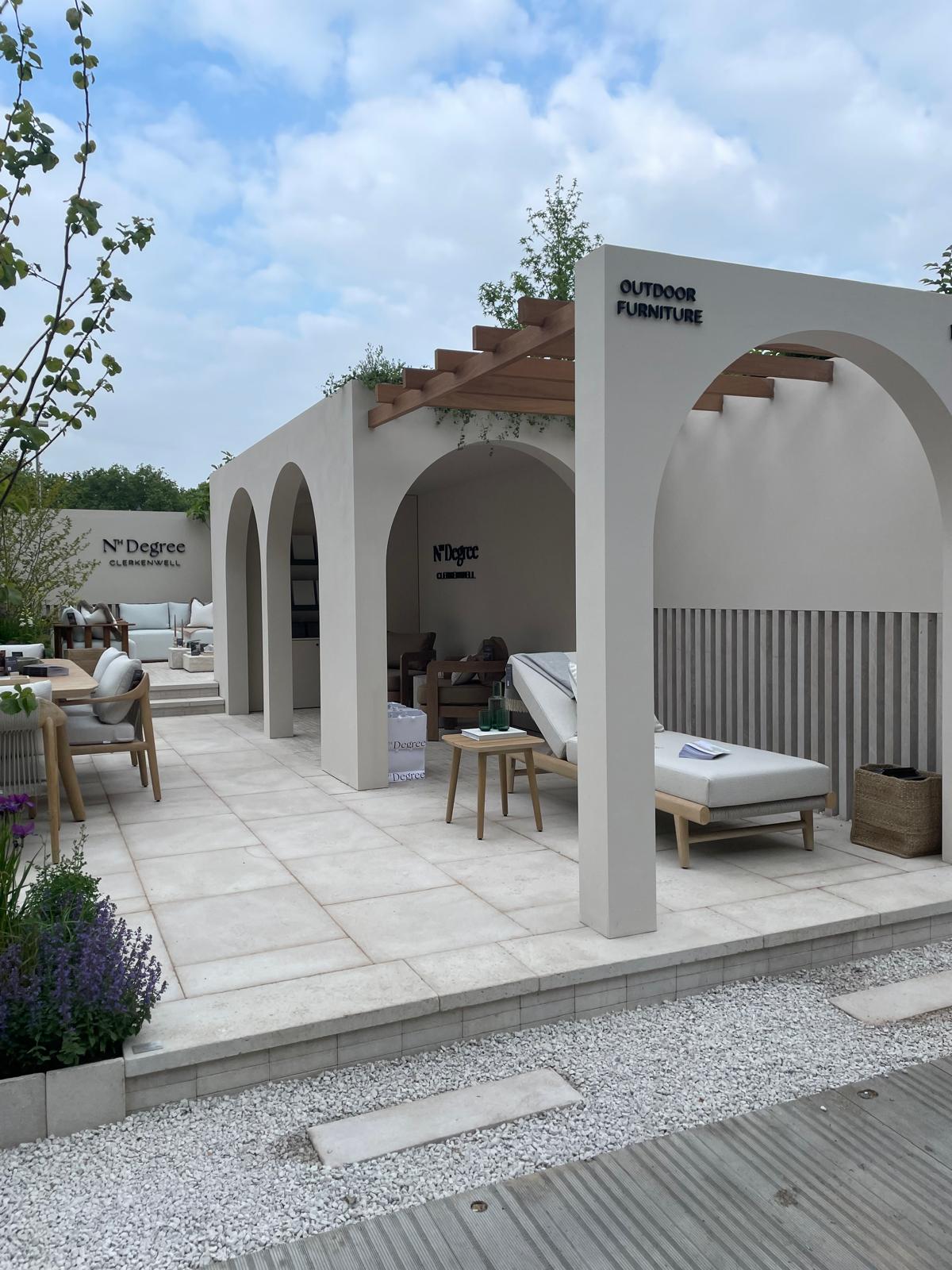 A modern outdoor space featuring lounge chairs under a pergola, surrounded by greenery and blue skies.