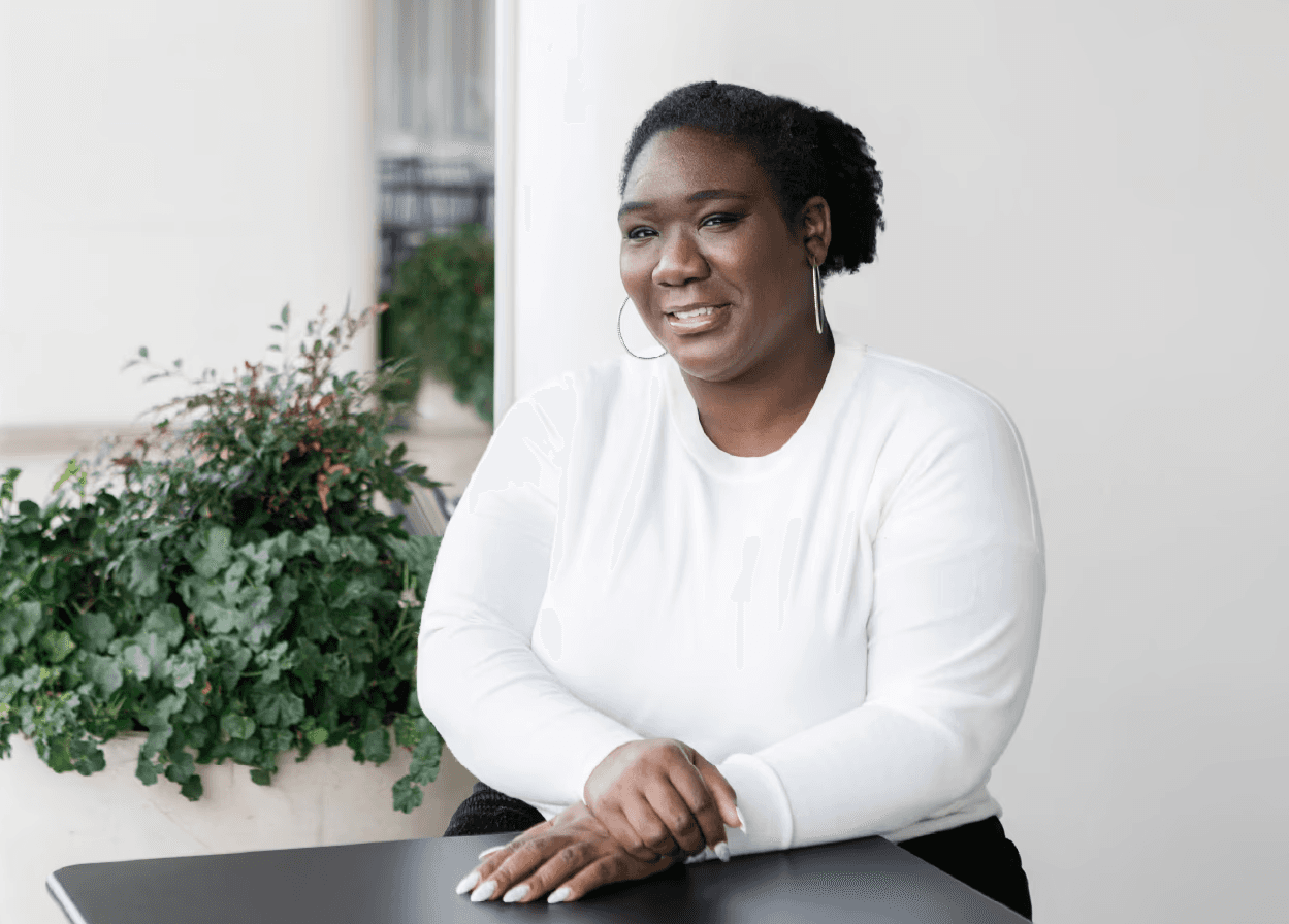 A medium shot captures therapist Sabrina Simpson with her hair pulled back, wearing a white long-sleeved shirt and l Sabrina Simpson is seated at a dark table, with her hands resting on its surface. Sabrina Simpson is looking to her right and smiling. Behind her, to the left, is a large planter filled with lush green foliage, and further back, a white pillar and a blurred background of more greenery and architectural elements. The lighting is soft and natural, creating a bright and airy atmosphere.