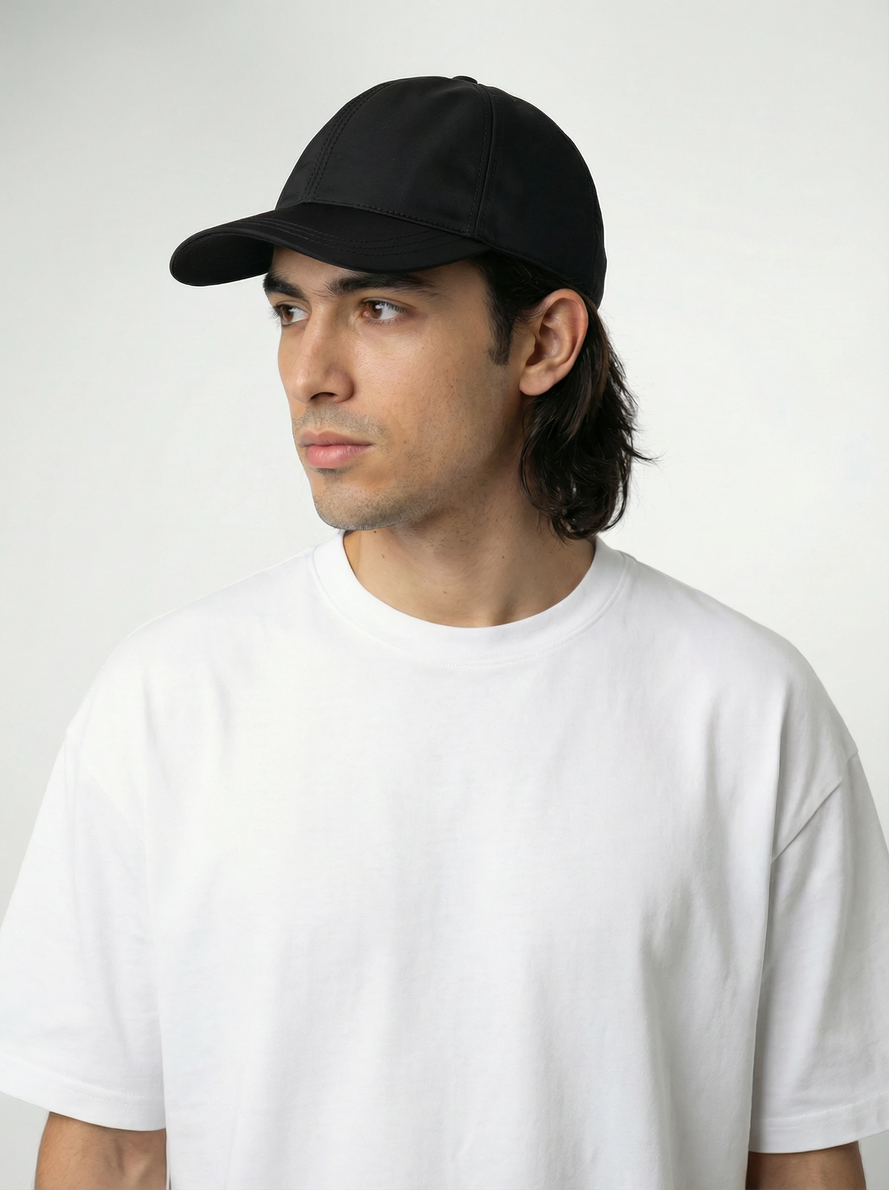 A person wearing a plain white t-shirt and a black baseball cap looks to the side, set against a neutral gray background.