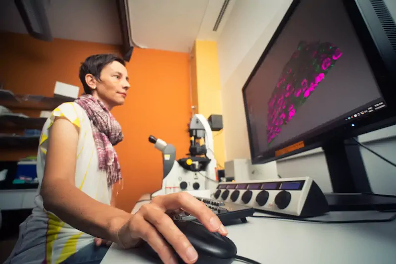 A female researcher operating a microscope in a lab, with a computer screen displaying biological imagery.