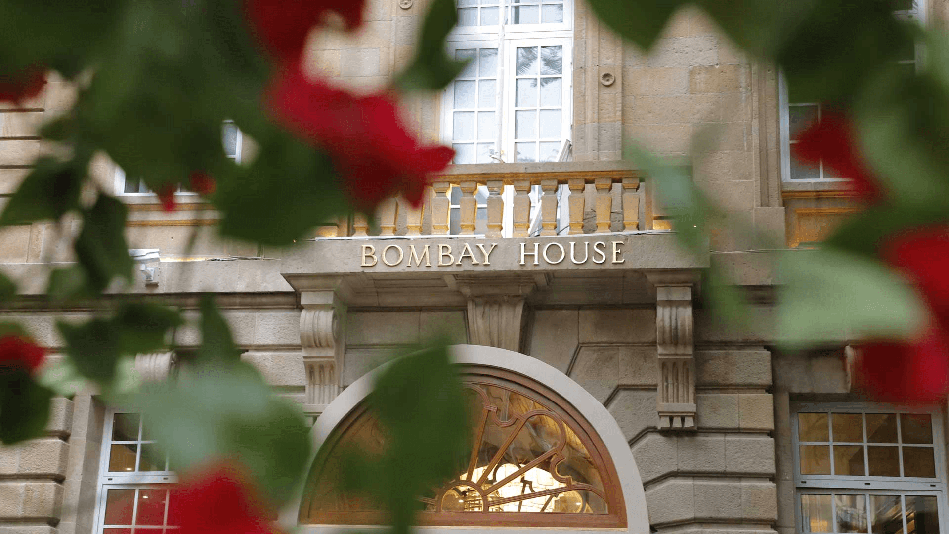 The facade of the historic Bombay House, framed by vibrant red flowers, features large, elegant windows and the building's name prominently displayed above a decorative archway.