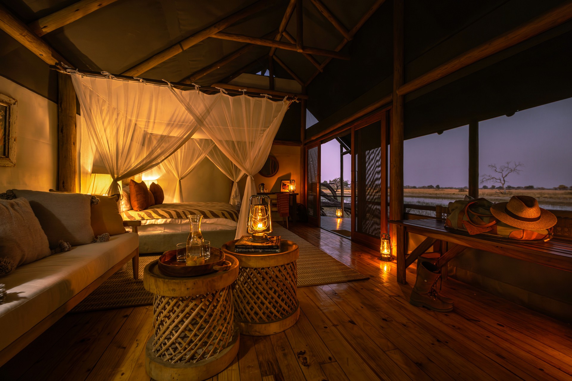 Kujwana Delta Ride, Okavango Delta, Botswana – Riverside Suites på Kujwana Camp, lyxigt boende med panoramavy över Okavango i kvällsljus. Vackert sken från lyktor och lampor belyser den stora dubbelsängen, vackert bäddad och omgiven av myggnät.