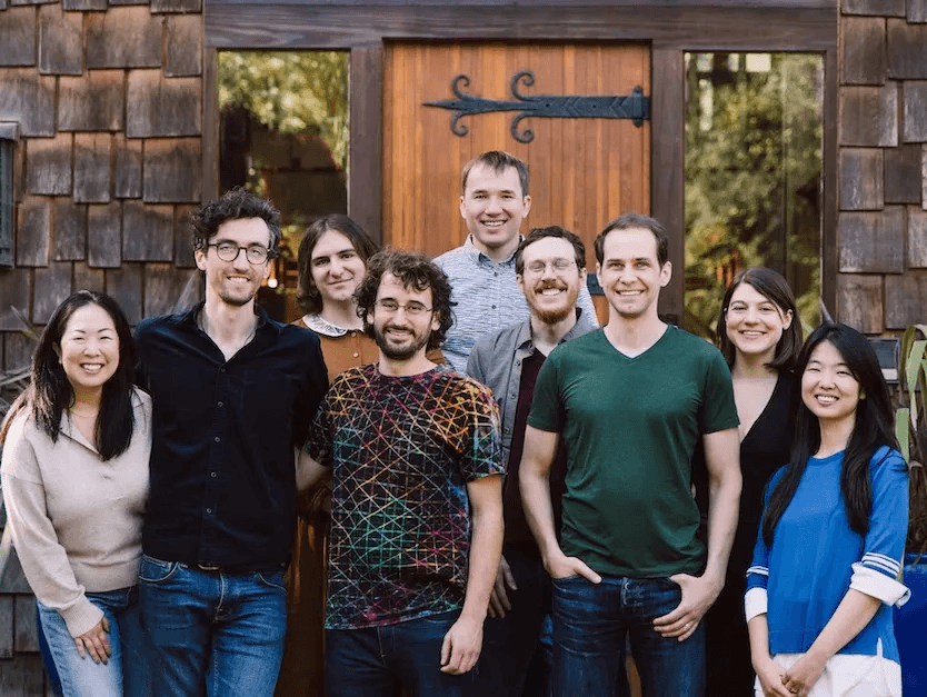 The Elicit team in early 2024. A group of nine people, casually dressed, smiling in front of a wooden door with rustic shingles, surrounded by greenery, conveys a friendly and relaxed gathering.