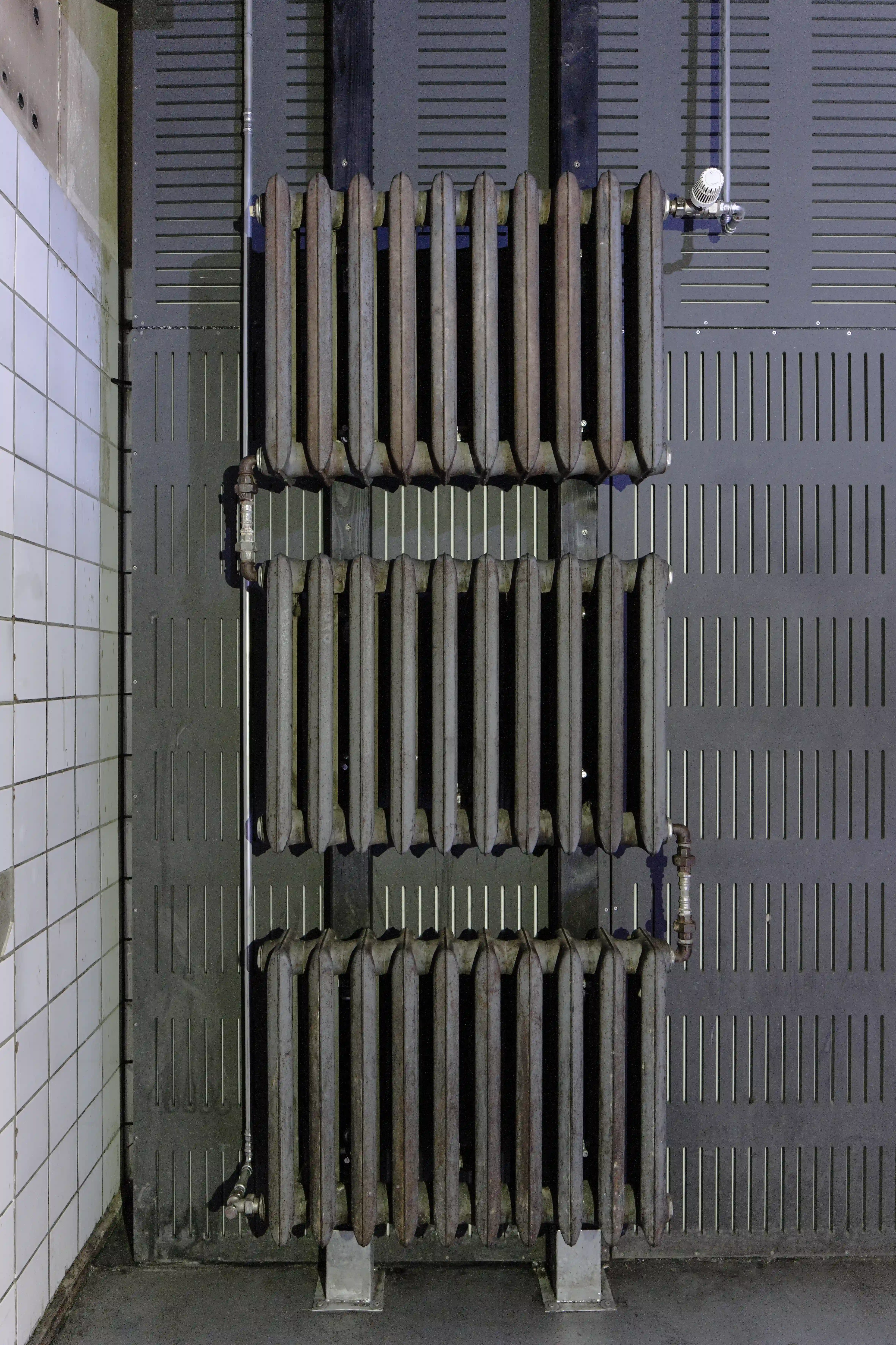 Detail at OHM CLUB of three vintage cast-iron radiators stacked vertically on a dark perforated metal wall next to white tiles.