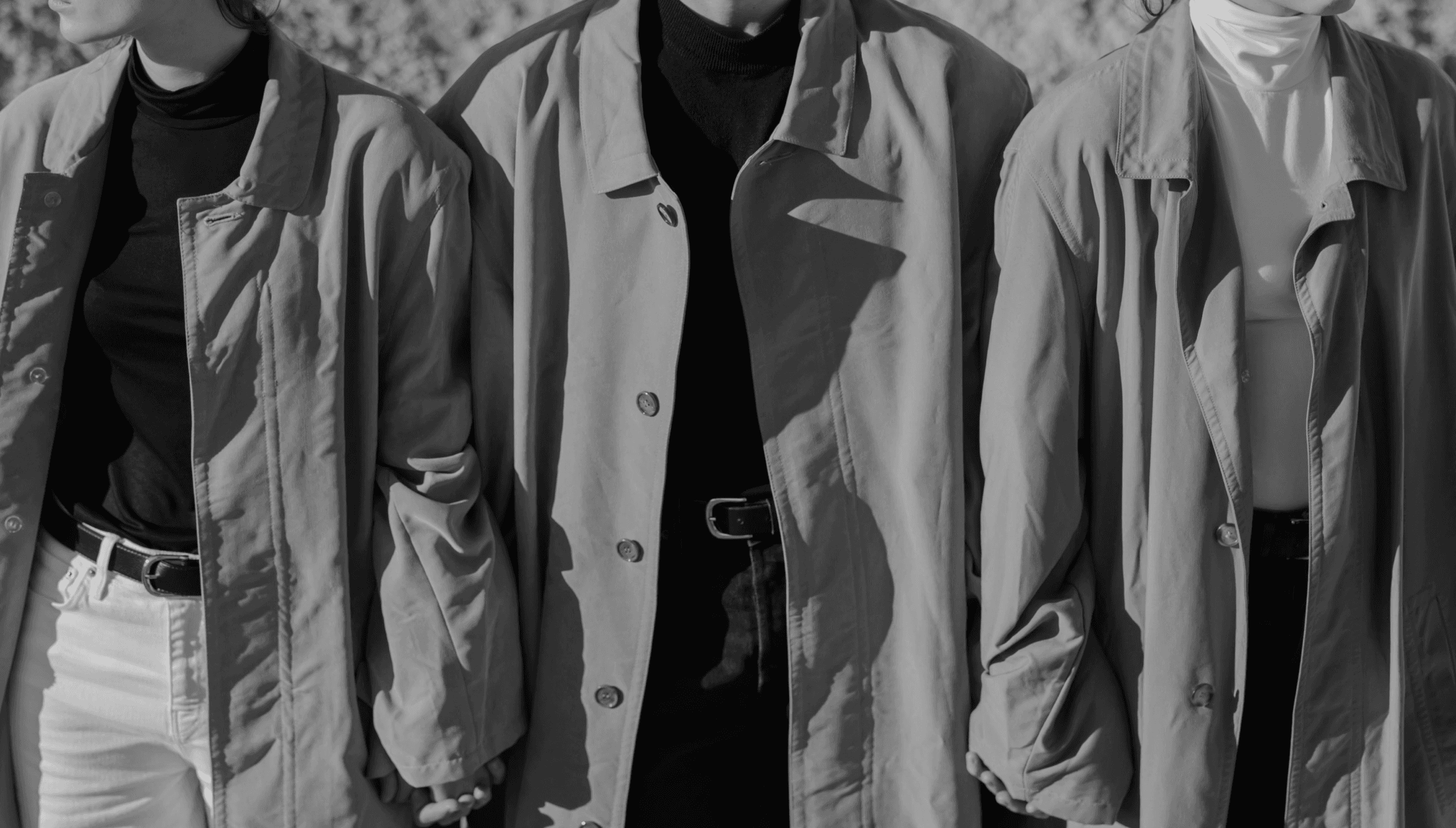 A tight, cropped view of three people standing side-by-side in trench coats, with the person in the middle wearing a dark turtleneck and a belted outfit.
