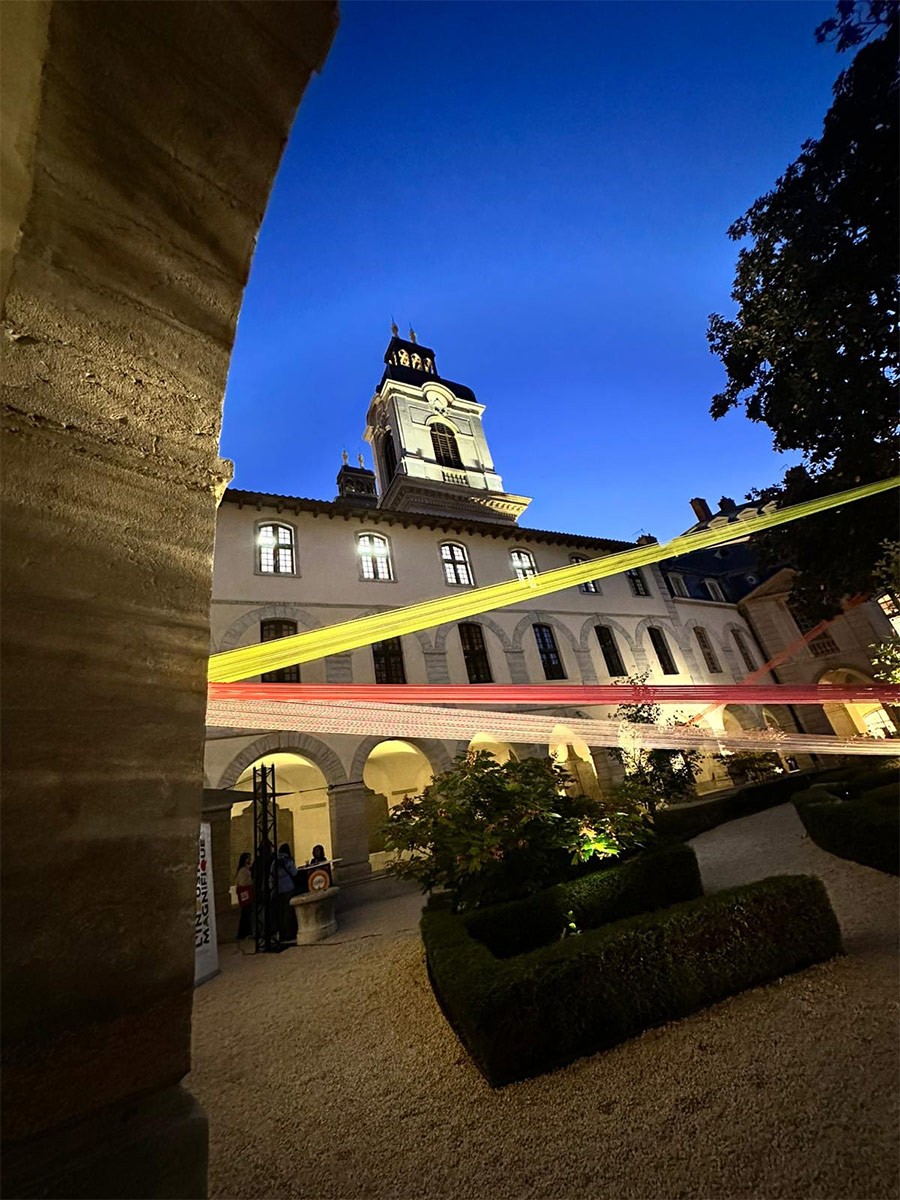 Une cour intérieure d'un bâtiment historique est illuminée par la lumière bleue du crépuscule. Des bandes de tissu colorées, jaune et rose, sont tendues en hauteur à travers la cour, reliant les façades du bâtiment qui présente des arcades et une tour centrale. Au premier plan, une allée en gravier et des haies taillées composent le jardin, tandis que quelques personnes sont visibles près de l'entrée du bâtiment sur la gauche.
