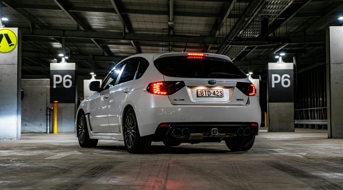 Une Subaru WRX blanche photographiée de dos dans un parking souterrain éclairé par des néons. Les quatre sorties d’échappement, l’assise large et l’éclairage arrière soulignent son tempérament sportif et son identité rallye assumée.
