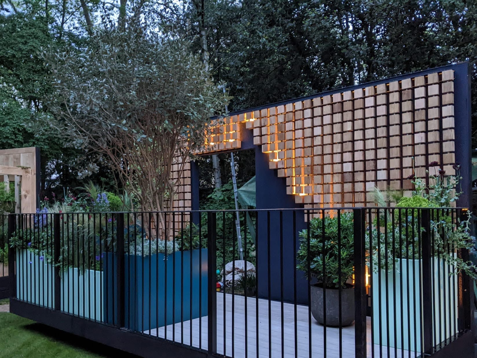 A modern outdoor space with wooden panels, plants, and a small blue area, surrounded by greenery.