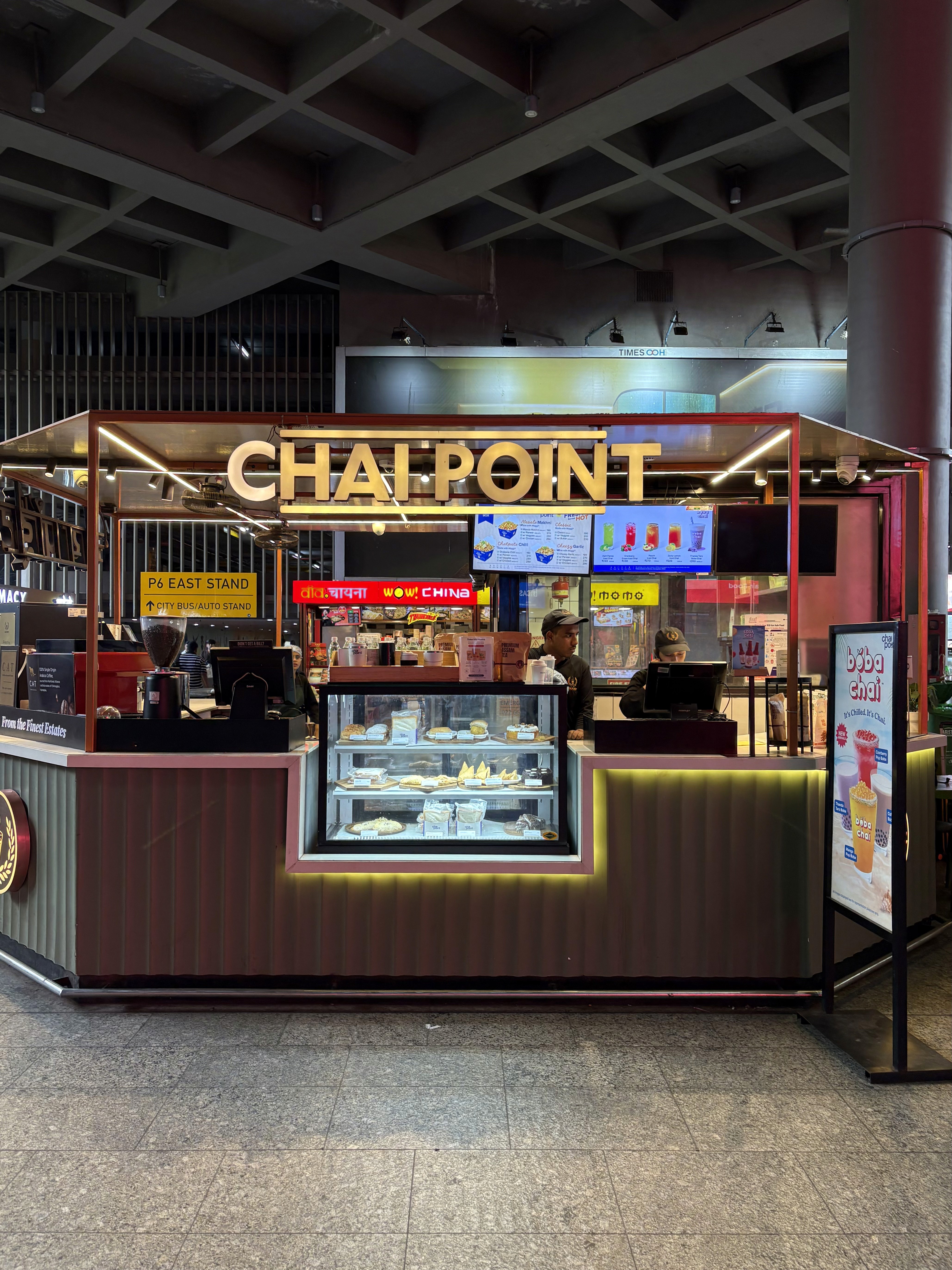 At Mumbai airport, a brightly lit Chai Point kiosk with a glowing sign, glass display of snacks, and digital menu boards. Staff are visible inside, serving customers in a busy indoor setting.