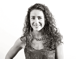 A black and white portrait of a young woman with long, curly hair. She is smiling and wearing a sleeveless top with a patterned design and a chain necklace. The background is plain white.