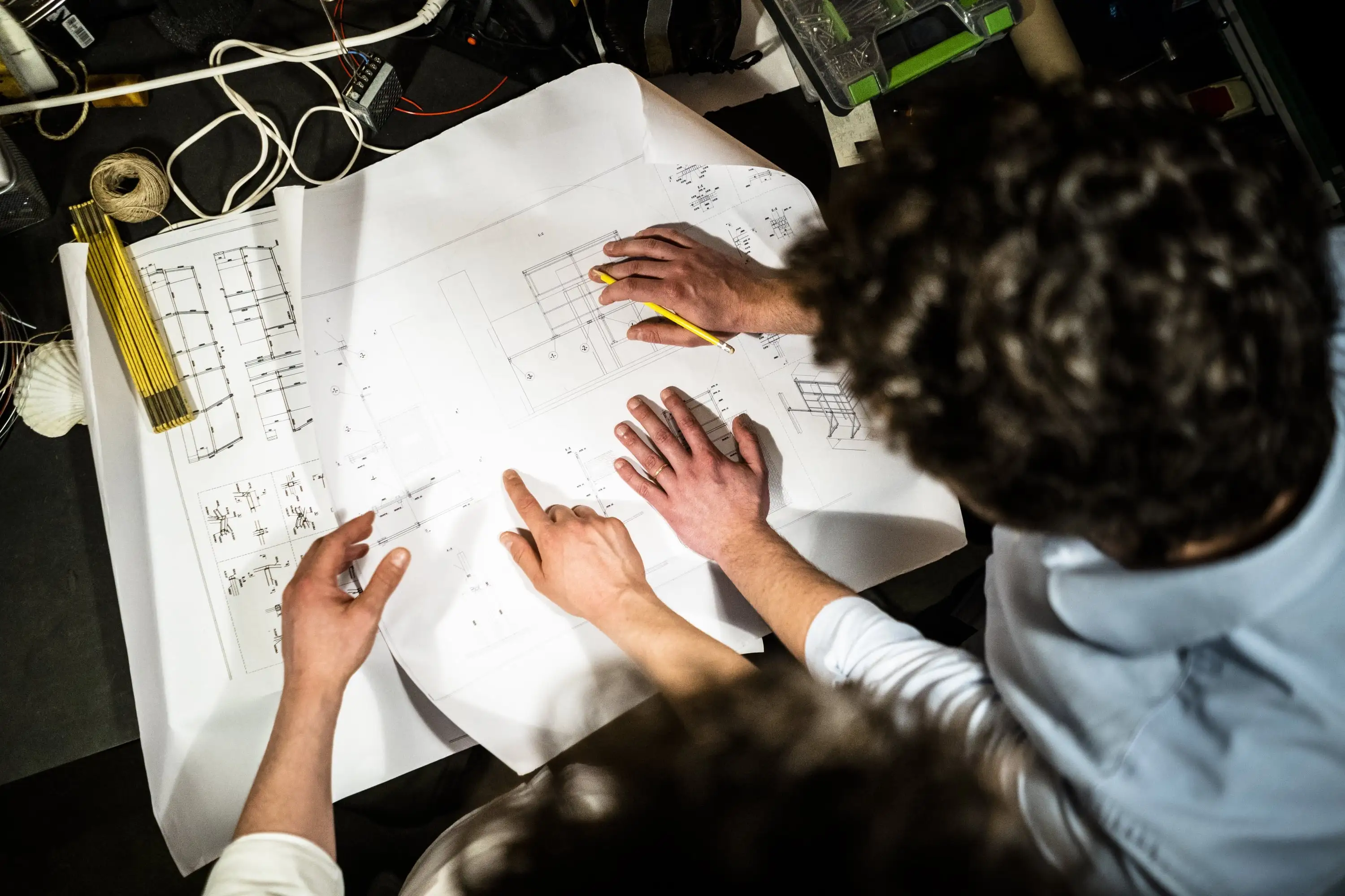 Zwei Personen überprüfen Baupläne auf einem Tisch mit Werkzeugen und Kabeln für ein Bauprojekt.