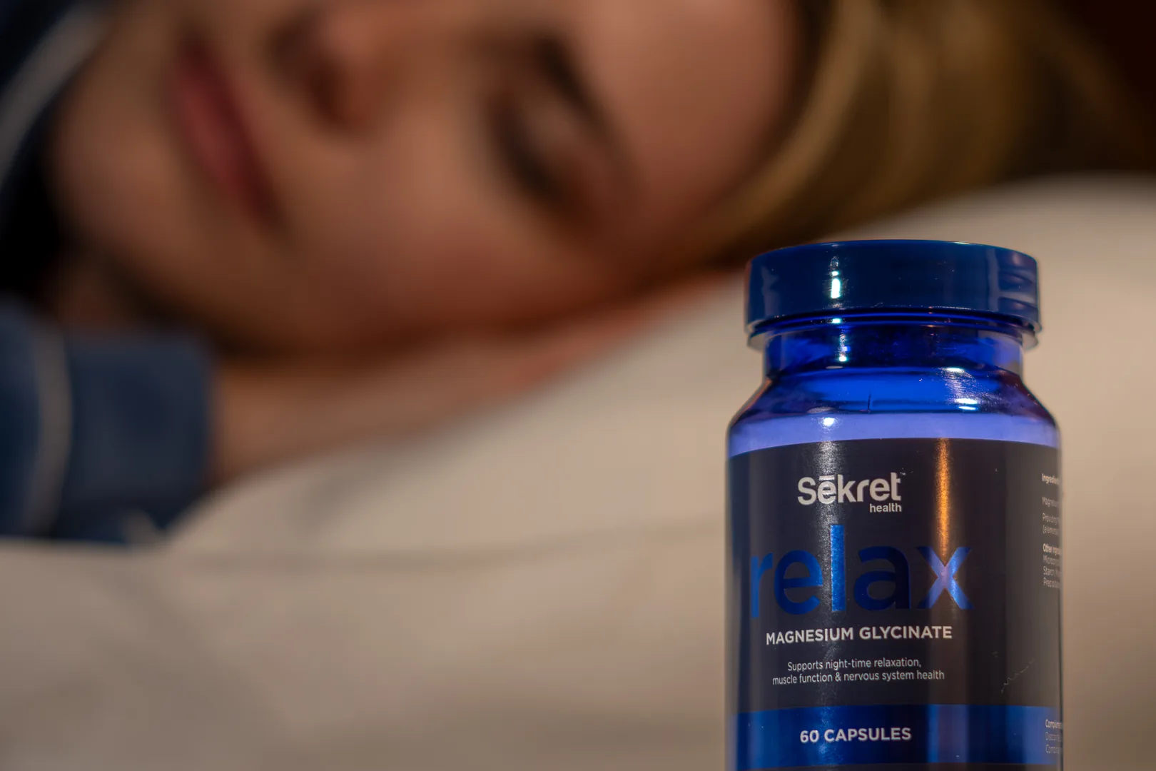 A bottle of Sēkret Relax capsules on a bedside table while a woman sleeps behind it
