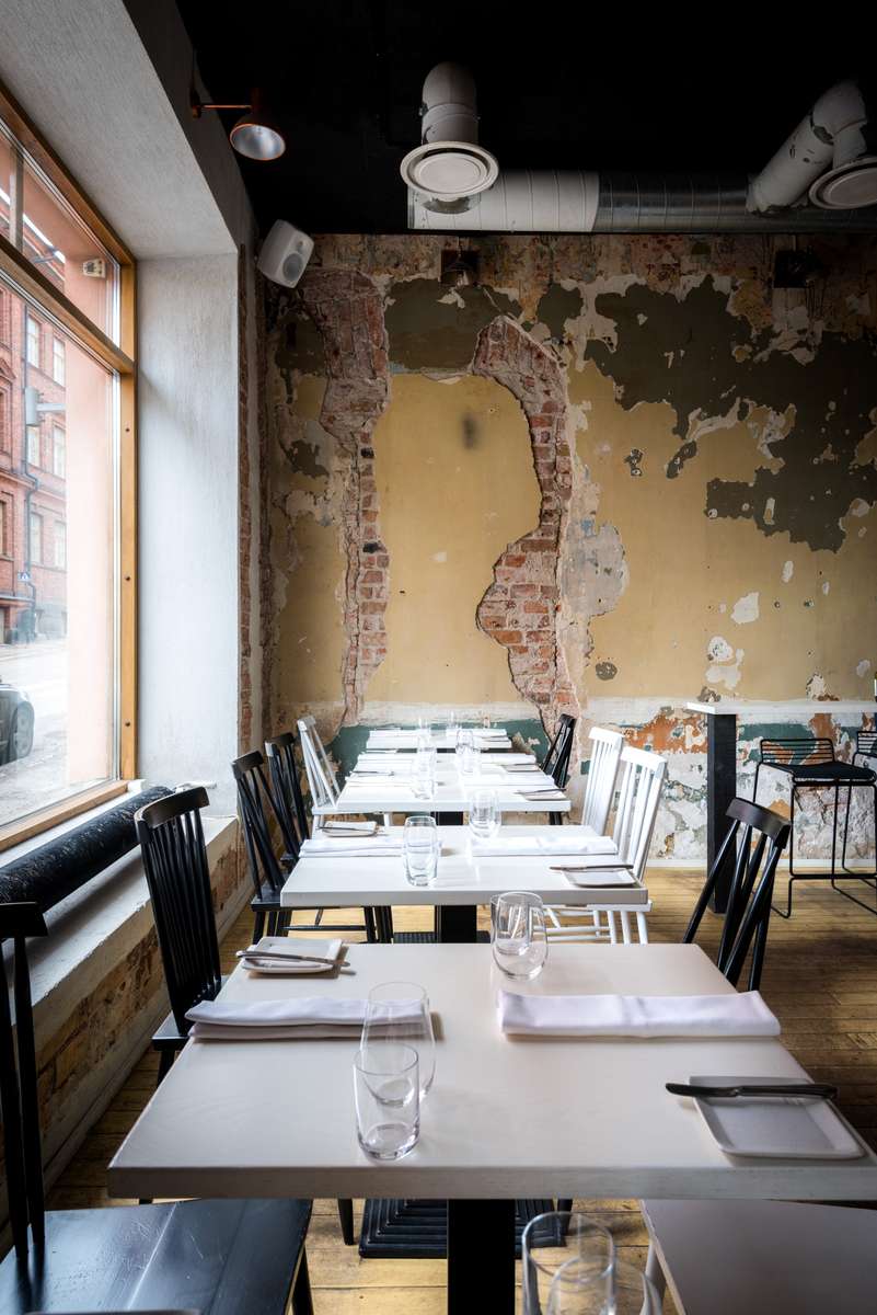 Rows of set tables and chairs line a narrow restaurant with an aged, peeling plaster wall and large windows.