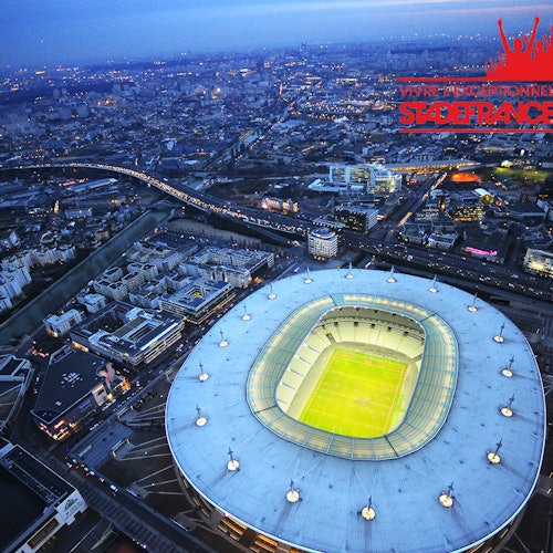 Aerial view of a brightly lit stadium in a bustling city at dusk with roads, buildings, and a river visible.