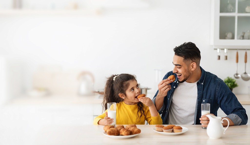 A smiling man and a young girl sit at a kitchen table enjoying pastries and glasses of milk together. To the left, a text banner reads “We help global brands expand their product lines in Saudi Arabia. We import, handle and place your products where people buy,” with yellow and white buttons labeled “Partner with us” and “See our brands,” and a WhatsApp icon in the lower corner.