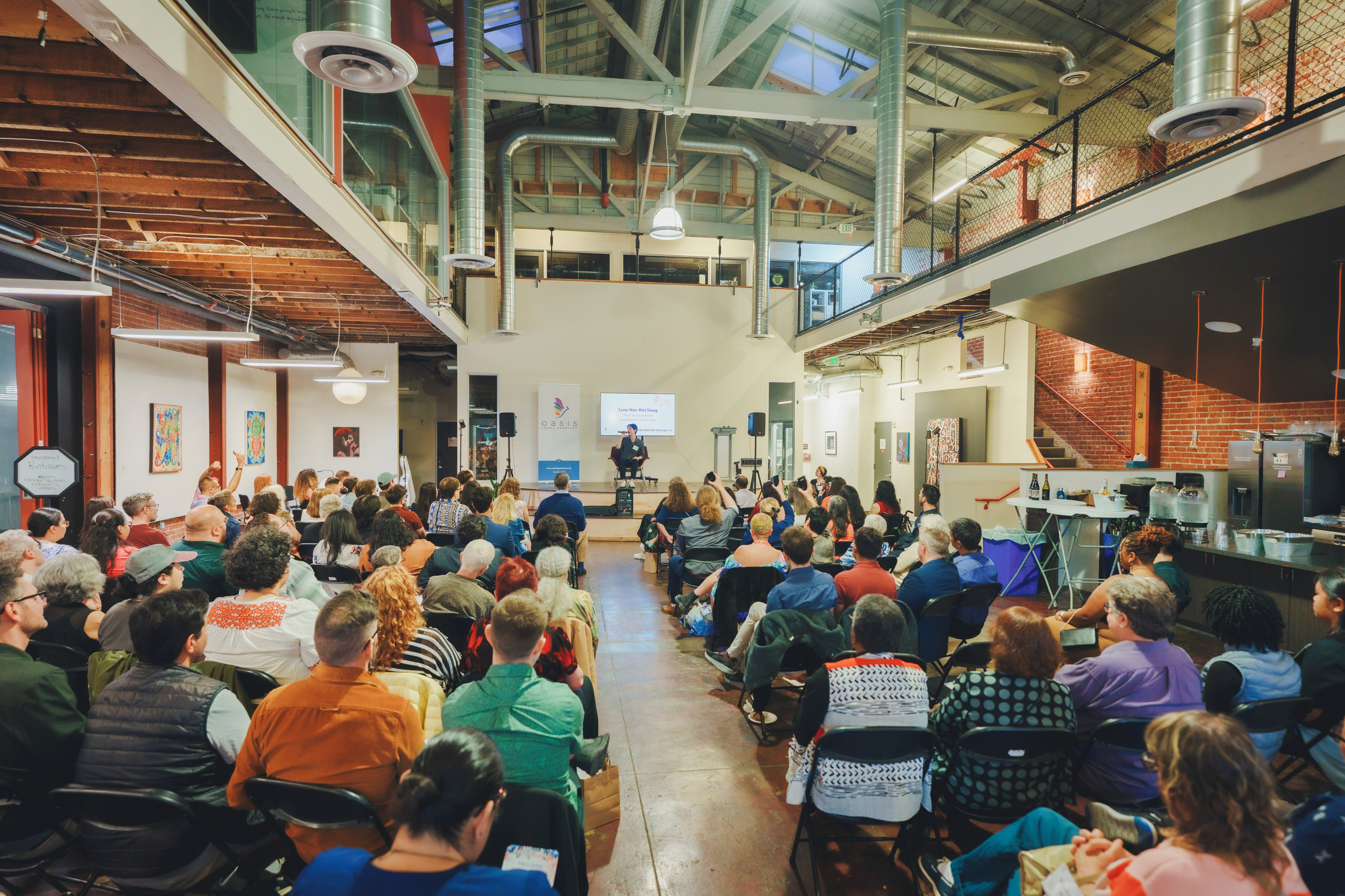 A diverse audience attentively listens to a speaker during a conference held in a spacious, industrial-style venue with exposed brick walls and high ceilings.
