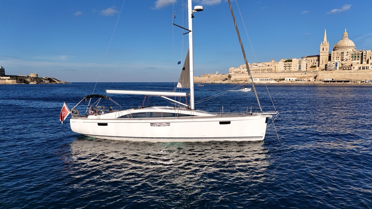 Beneteau Clipper 361 sailing yacht motoring at anchor in calm blue waters off Valletta, Malta, showing the full port profile with white hull, furled mainsail, bimini with solar panel, B&G instruments on the stern arch, and the iconic Valletta skyline with St Paul's Cathedral dome and historic limestone fortifications behind