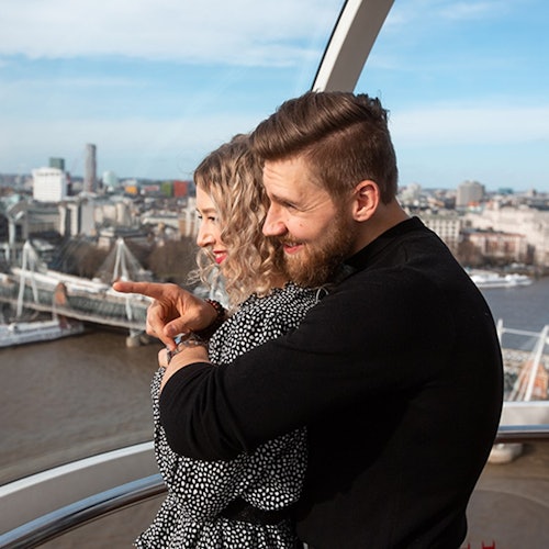 A couple embraces inside a ferris wheel pod, overlooking a river and a cityscape with bridges and buildings in the background.