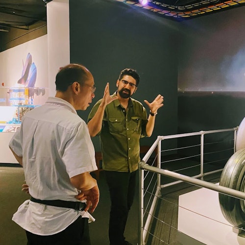 Two men are conversing in an exhibition space, one gesturing expressively. An industrial object is partially visible in the foreground.