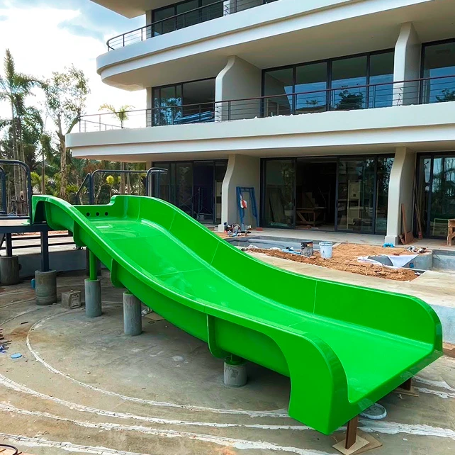 Cầu trượt màu xanh lá cây tươi sáng được đặt phía trước tòa nhà hiện đại, nhiều tầng đang được xây dựng, với cây cối xung quanh và bầu trời trong xanh ở phía sau.