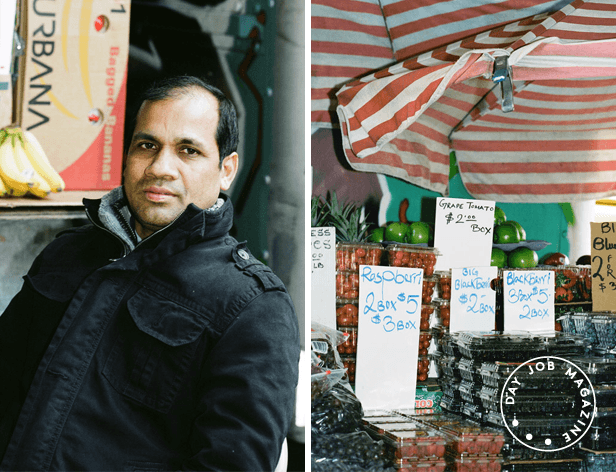 A man with short dark hair and a dark jacket stands on the left side of the image, facing forward. Behind him, a box labeled “Bagdad Banana” is visible, along with a bunch of ripe bananas. To the right, a fruit stand displays signs for grape tomatoes and raspberries, with price tags showing “$2.00 Box” and “$3.00 Box”. The stand also has containers of blackberries and other fruits. A red-and-white striped awning covers the stand.