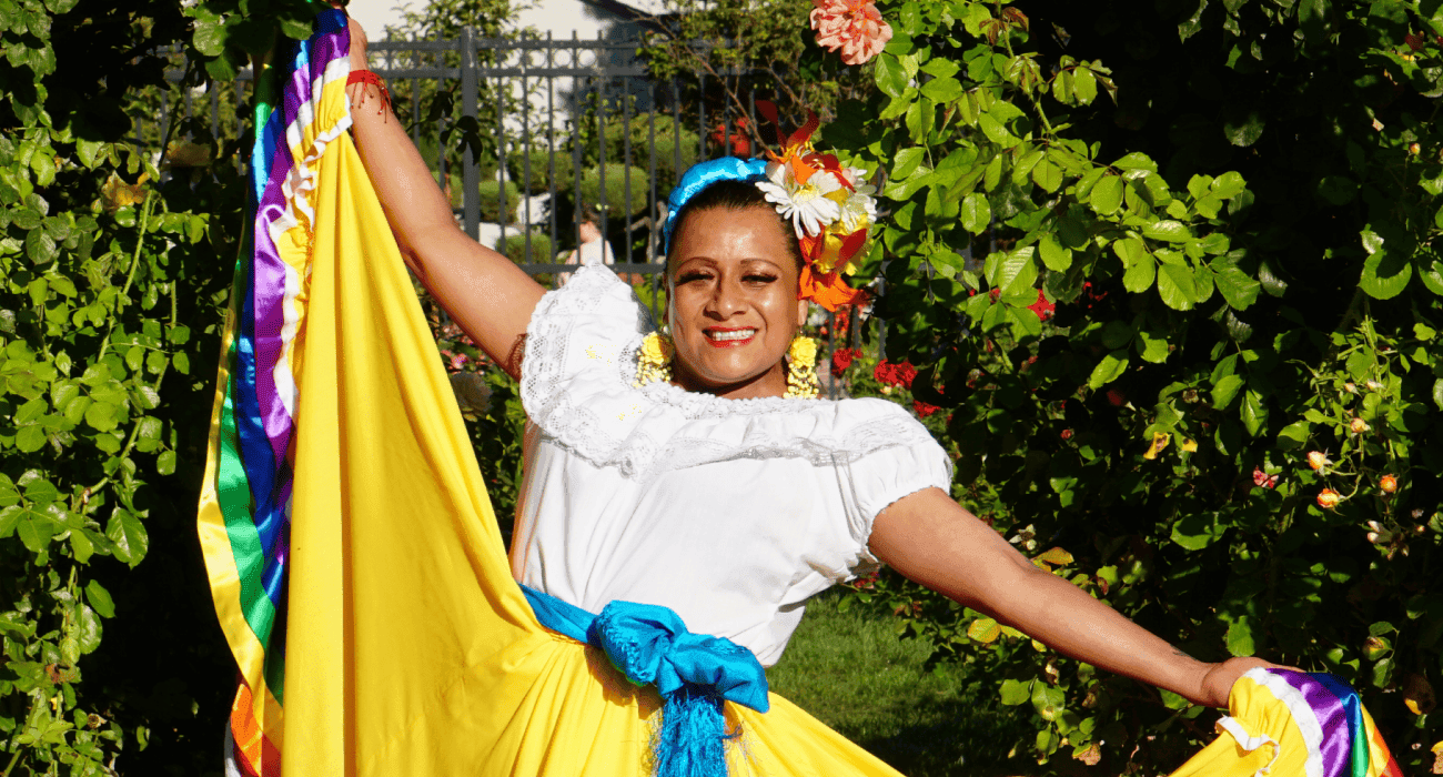 Una persona con atuendo tradicional de danza folclórica mexicana posa alegremente en un jardín, luciendo una vibrante falda amarilla con un dobladillo colorido y una blusa blanca adornada con brillantes accesorios florales y una banda azul.