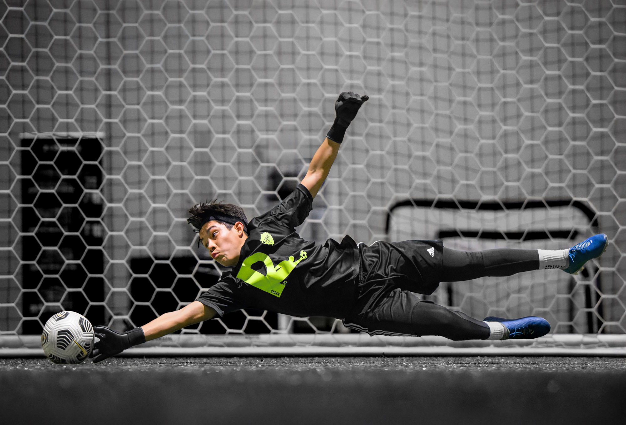 A youth goalkeeper dives horizontally, extending his right arm to save a soccer ball in front of the net.