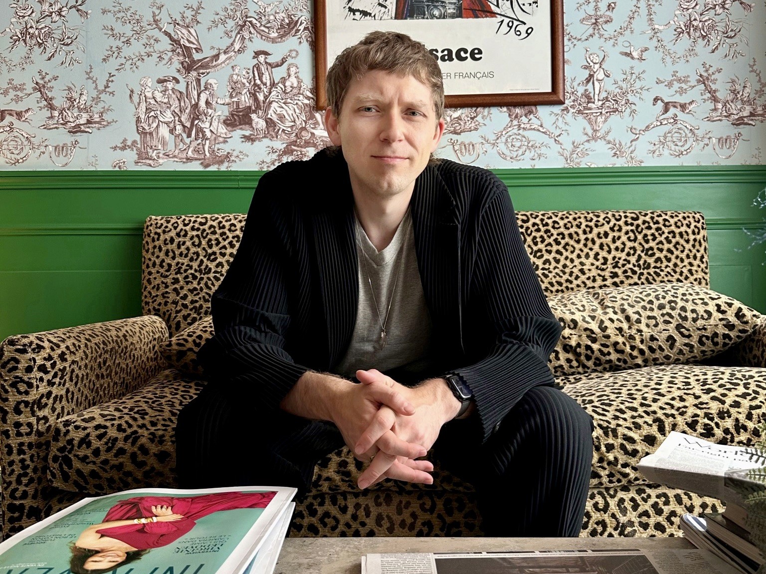Editorial director and strategist JD Shadel leans against a wall and looks off into the distance. They have shaggy blonde hair and wear a vintage leather jacket over a roll neck sweater. 