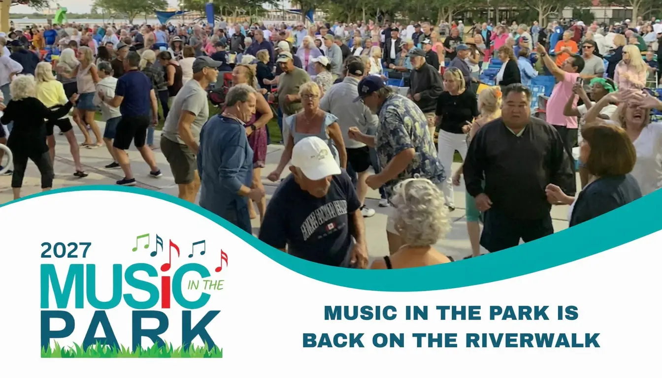 A lively crowd gathers at the 2027 Music in the Park event on the Riverwalk in Bradenton, FL, with people dancing and enjoying the live music under a clear sky.