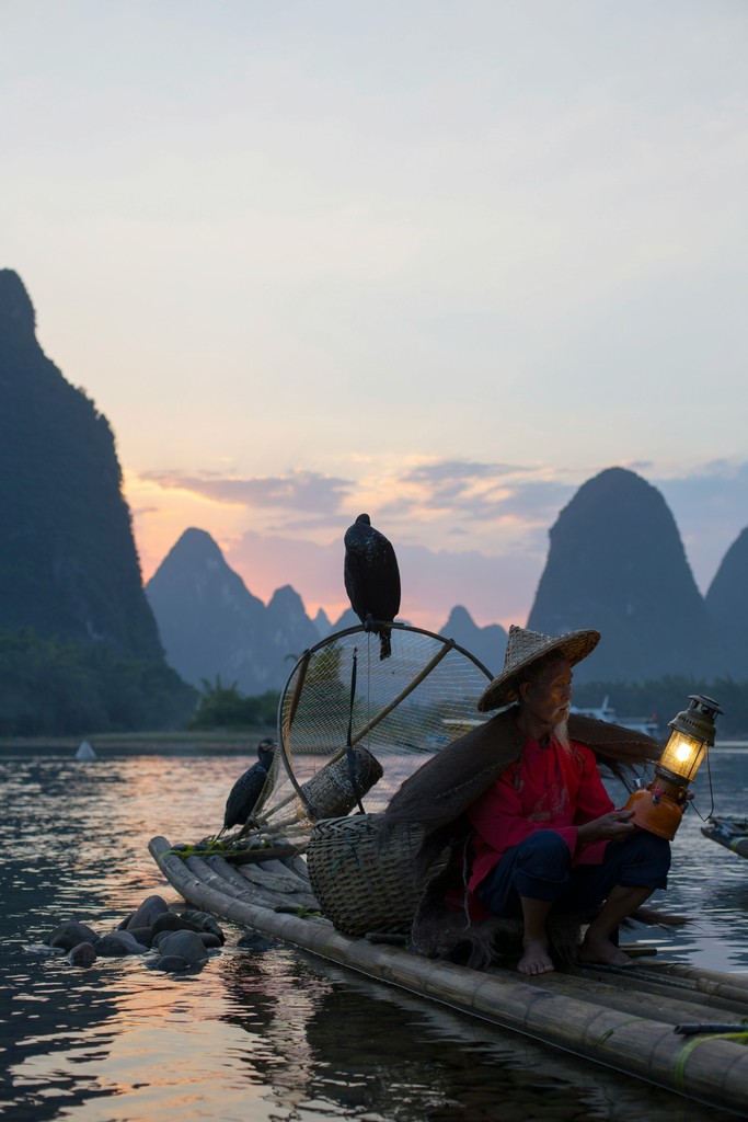 An elderly fisherman wearing traditional clothing and a straw hat holds a lantern on a bamboo raft, accompanied by cormorants, set against the backdrop of the picturesque Karst mountains at sunset.