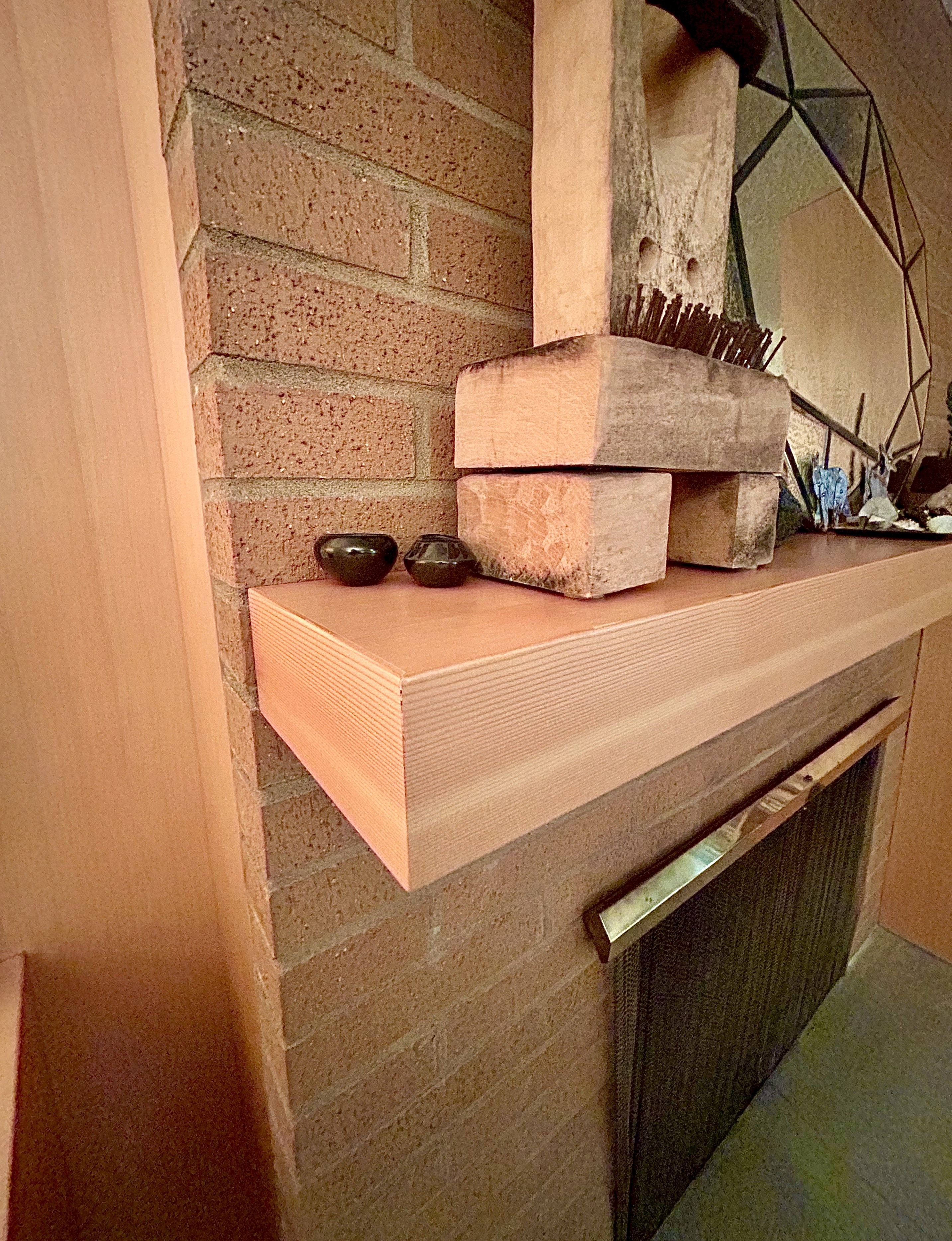 A thick wooden mantle in front of a brick wall and over a fireplace holds art and a mirror.
