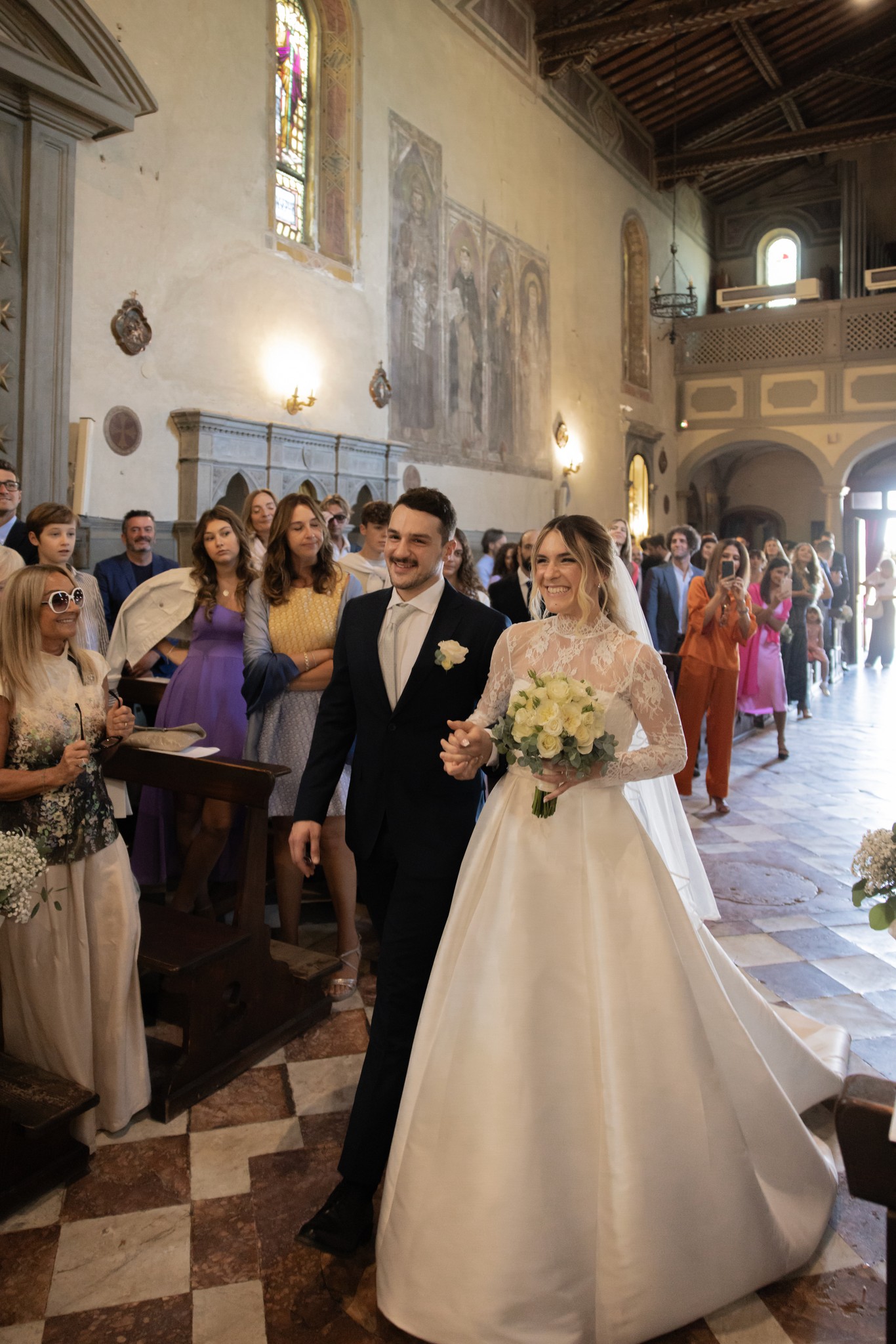 Sposi in navata durante cerimonia a Bolgheri, lei in abito bianco con bouquet, lui in smoking, ospiti ai lati